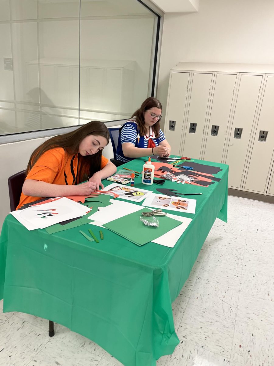 Spooky crafts. Waiting for students and the little kids to arrive, junior Amelia Watson [left] and junior Mila Hormann [right]  volunteer with Art Club for Bellevue East’s BEAST Trick or Treat Night. They were in charge of running stations around the school in which kids stopped by and glued together Halloween themed characters such as pumpkins, ghosts, and Frankensteins. The Trick or Treat night is a fun opportunity for kids to connect with high schoolers and play games while dressed up in costumes. “BEAST Trick or Treat has been my favorite activity," Watson said. “It was fun working with little kids.”